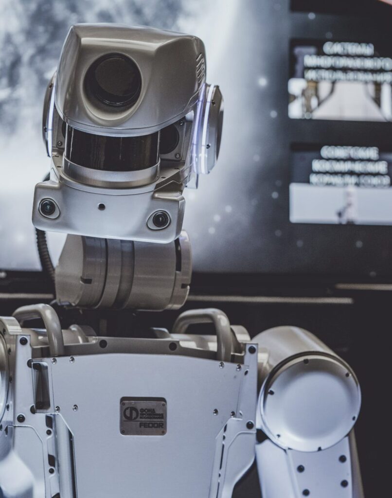 Ein humanoider Roboter mit einem metallischen silbernen Körper ist im Bild prominent zu sehen. Der Roboter hat einen großen, quadratischen Kopf mit einer zentralen, schwarzen kreisförmigen Linse, die einem Auge ähnelt und ihm ein futuristisches Aussehen verleiht. Auf beiden Seiten des Kopfes befinden sich runde, beleuchtete Komponenten, die möglicherweise Sensoren oder Lichter sind. Der Hals des Roboters besteht aus einer Reihe von metallischen Segmenten, die Bewegungen ermöglichen. Sein Torso ist schlank und eckig, mit sichtbaren Schrauben und Paneelen, und hat ein robustes, industrielles Design. Auf der Brust befindet sich eine Metallplatte mit der eingravierten Aufschrift "ФОНД НАЦИОНАЛЬНЫХ ПРОЕКТОВ" und "FEDOR". Im Hintergrund ist ein verschwommener Bildschirm zu sehen, der einige Texte und Bilder anzeigt, was auf eine technologische oder Ausstellungssituation hindeutet. Die Gesamtatmosphäre ist eine von fortschrittlicher Robotik und Innovation.
