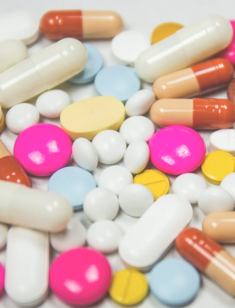Eine Nahaufnahme einer Vielzahl von Tabletten und Kapseln, die auf einer weißen Oberfläche verstreut sind. Die Auswahl umfasst runde, ovale und kapselartige Pillen. Die Farben sind vielfältig und umfassen leuchtendes Pink, Hellblau, Weiß, Gelb und eine zweifarbige Kapsel in Beige und Orange. Einige Pillen sind glänzend und glatt, während andere eine matte Oberfläche haben. Die pinkfarbenen Pillen sind rund und glänzend, die blauen sind flach und rund, die weißen Pillen sind sowohl rund als auch oval, und die gelben sind klein und rund mit einer Linie in der Mitte. Die beige und orangefarbenen Kapseln sind länglich mit einer glänzenden Oberfläche. Die Anordnung ist insgesamt zufällig, was eine lebendige und farbenfrohe Darstellung ergibt.