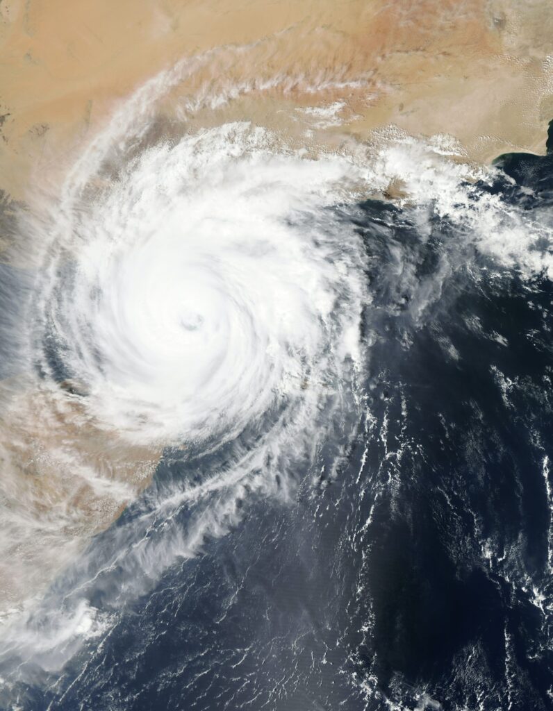 Ein Satellitenbild zeigt einen massiven Zyklon über dem Arabischen Meer, in der Nähe der Küste von Oman. Der Zyklon hat ein deutlich erkennbares, kreisförmiges Auge in seinem Zentrum, umgeben von eng gewundenen Bändern aus dichten, weißen Wolken. Die Wolken erstrecken sich in einer gegen den Uhrzeigersinn gerichteten Richtung, was auf die Rotation des Sturms hinweist. Der Ozean unter dem Zyklon erscheint dunkelblau und steht im scharfen Kontrast zum strahlenden Weiß der Wolken. Im oberen Teil des Bildes ist das Festland von Oman sichtbar, gekennzeichnet durch seine sandige, wüstenähnliche Landschaft in hellbraunen und beigen Farbtönen. Die Küstenlinie ist unregelmäßig, mit einigen Bereichen, die ins Meer ragen. Das Bild erfasst die immense Größe und Kraft des Zyklons, während er sich dem Land nähert.