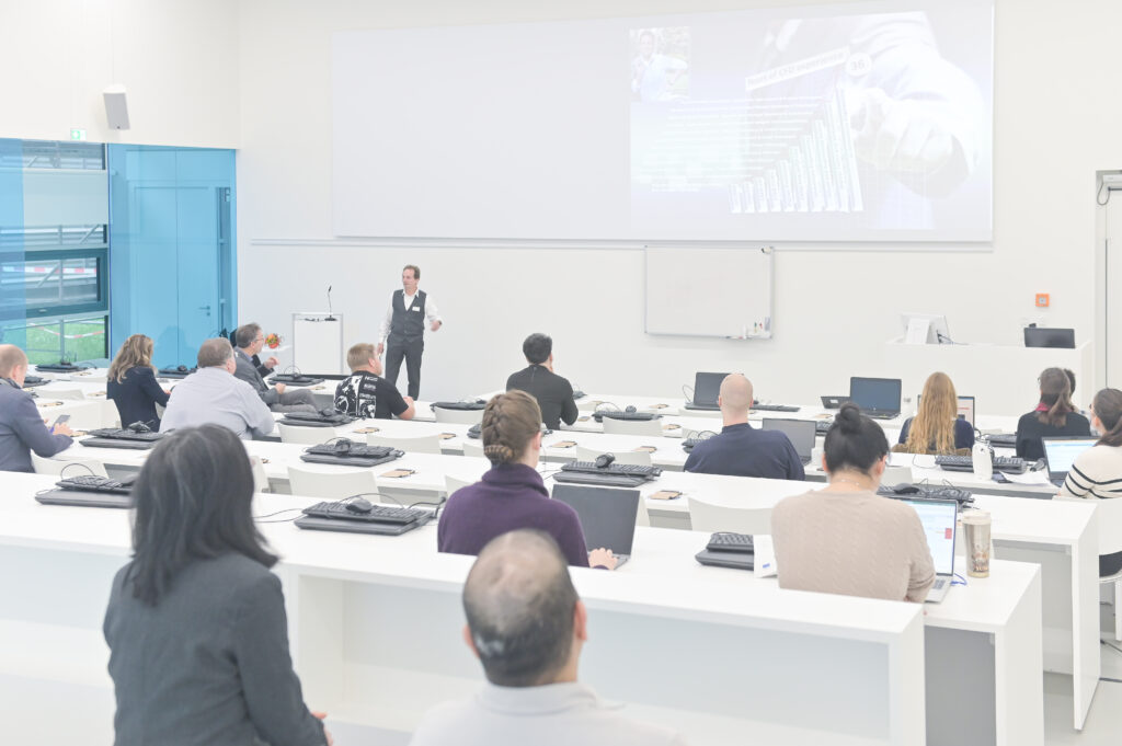 Ein Referent präsentiert vor einem aufmerksam zuhörenden Publikum im modernen, lichtdurchfluteten Hörsaal des HLRS. Auf der Leinwand ist eine Präsentationsfolie mit Diagrammen zu sehen.
