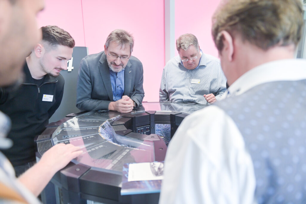 Vier Männer stehen um einen interaktiven Bildschirm herum und schauen auf die beleuchtete Oberfläche, auf der digitale Inhalte angezeigt werden. Die beiden Männer im Vordergrund stehen mit dem Rücken zum Betrachter, während die beiden Männer in der Mitte, die eine Brille tragen, konzentriert auf den Bildschirm schauen.