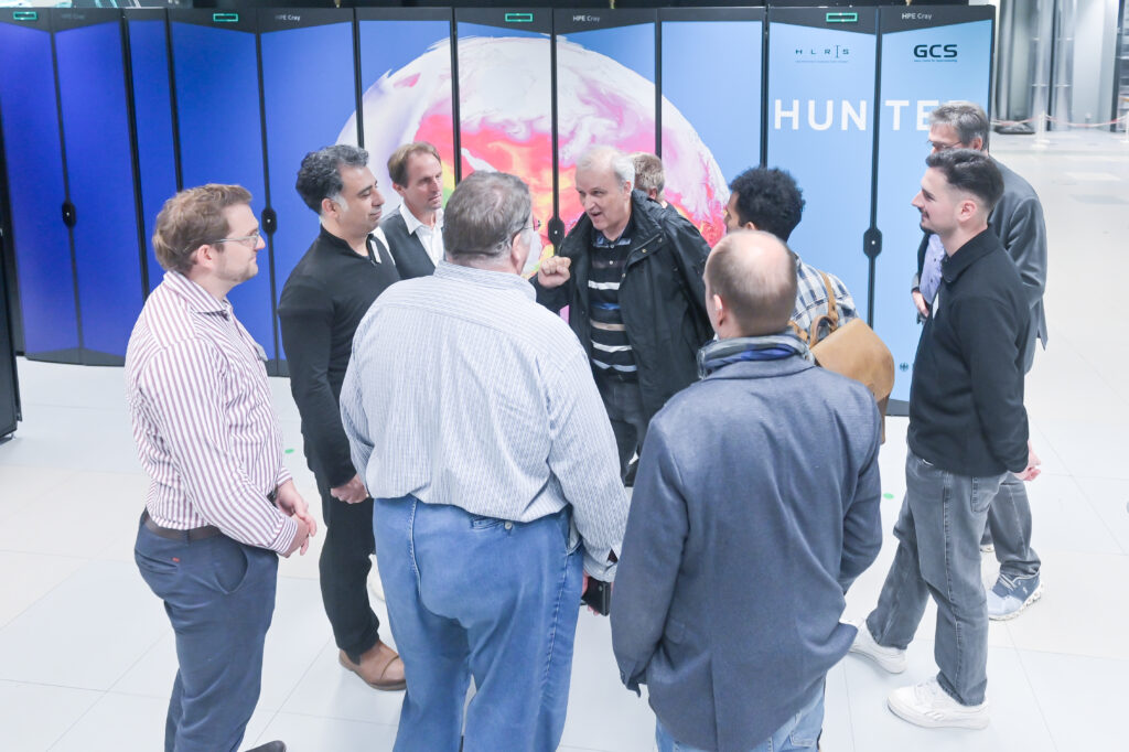 Eine Gruppe von neun Personen, hauptsächlich Männer, hat sich im HLRS-Rechenzentrum versammelt, im Hintergrund ist Hunter zu sehen. Sie sind während der Führung mit Hunter in eine Diskussion vertieft, wobei ein älterer Mann in einer dunklen Jacke gestikulierend zu der Gruppe spricht.