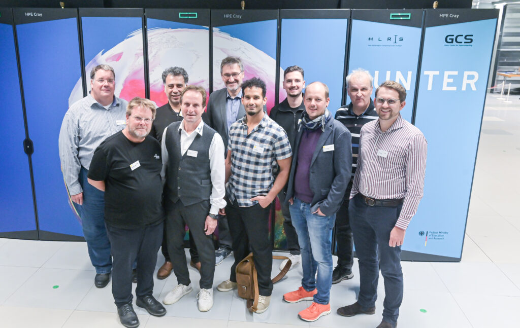 Zehn Männer posieren für ein Gruppenfoto in einem Raum mit großen blau-weißen Computerserver-Racks im Hintergrund, die mit „HPE Cray” und „HLRS GCS HUNTER” beschriftet sind. Sie blicken alle in die Kamera und lächeln, gekleidet in einer Mischung aus Freizeit- und Business-Casual-Kleidung.