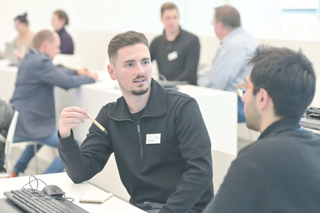 Ein junger Mann mit Bart und dunklem Pullover mit Viertelreißverschluss sitzt an einem weißen Tisch, hält einen Bleistift in der Hand und schaut zu jemandem außerhalb des Bildausschnitts zu seiner Rechten. Im unscharfen Hintergrund sitzen weitere Personen an ähnlichen Tischen, unterhalten sich oder arbeiten.