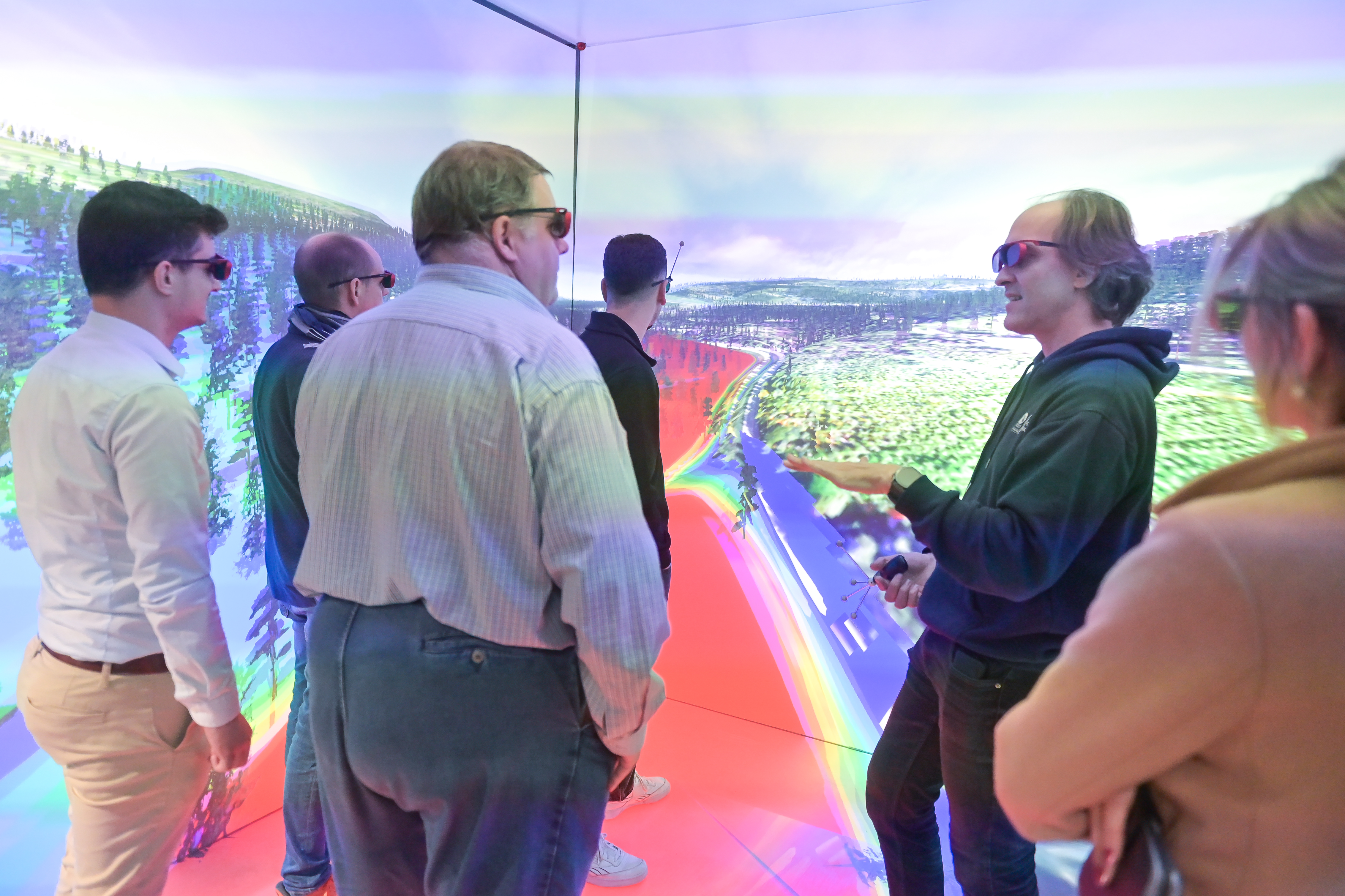 Eine Gruppe von fünf Personen, die alle 3D-Brillen tragen, steht in einem immersiven Virtual-Reality-Raum, dessen Wände eine farbenfrohe, dynamische Landschaft zeigen. Ein Mann auf der rechten Seite, der einen dunklen Kapuzenpulli trägt, gestikuliert mit den Händen, als würde er den anderen, die die projizierte Umgebung betrachten, etwas erklären.